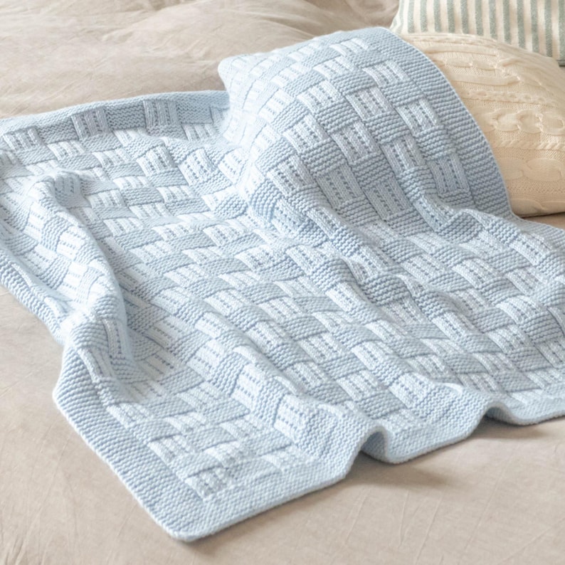 May include: A light blue knitted blanket and pillow set with a woven basket weave pattern. The blanket is folded over a bed, with a matching pillow resting on top. The blanket has a thick border.