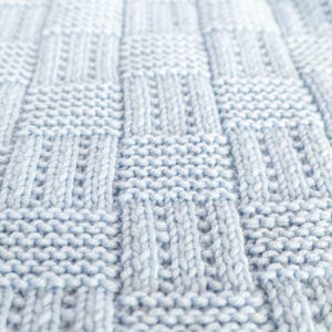 May include: Close-up of a light blue knitted blanket with a textured, woven pattern. The blanket's design features a series of rectangular and vertical knit stitches, creating a tactile and visually appealing surface. The soft, pastel color suggests comfort and warmth.