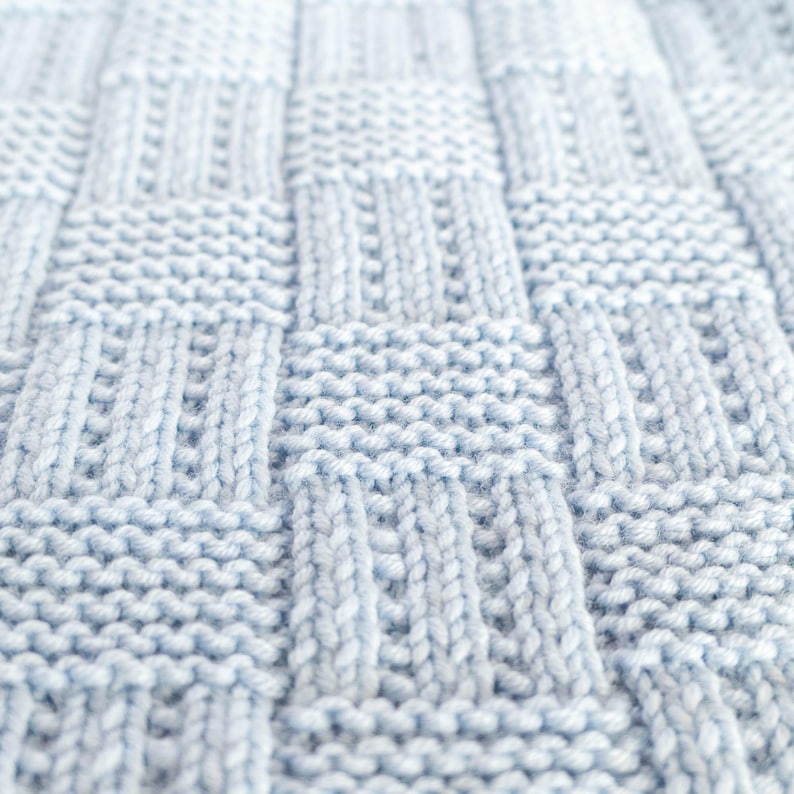 May include: Close-up of a light blue knitted blanket with a textured, woven pattern. The blanket's design features a series of rectangular and vertical knit stitches, creating a tactile and visually appealing surface. The soft, pastel color suggests comfort and warmth.