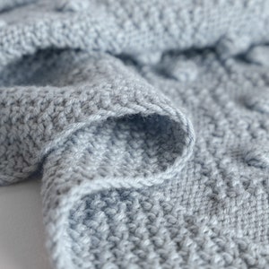 Pu&ograve; includere: Primo piano di una morbida coperta lavorata a maglia azzurra. La coperta presenta un motivo a punto strutturato con bobi in rilievo, creando un design tattile e visivamente accattivante. I bordi sono rifiniti con cura, mostrando la qualit&agrave; dell'artigianato.