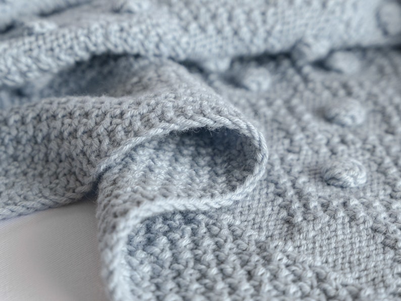 Pu&ograve; includere: Primo piano di una morbida coperta lavorata a maglia azzurra. La coperta presenta un motivo a punto strutturato con bobi in rilievo, creando un design tattile e visivamente accattivante. I bordi sono rifiniti con cura, mostrando la qualit&agrave; dell'artigianato.
