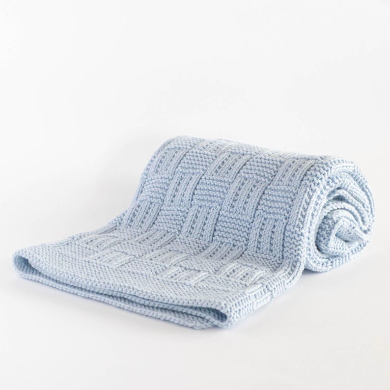 May include: A light blue knitted blanket with a woven basket weave pattern. The blanket is rolled up on one end and lies flat on a white surface. The blanket is made of soft material.
