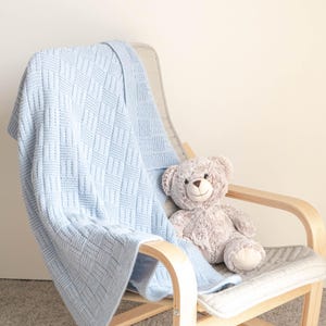 May include: A light blue knitted baby blanket with a geometric pattern is draped over a wooden chair. A plush teddy bear sits on the chair. The blanket is made of soft material and is suitable for a nursery.