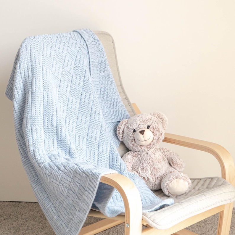 May include: A light blue knitted baby blanket with a geometric pattern is draped over a wooden chair. A plush teddy bear sits on the chair. The blanket is made of soft material and is suitable for a nursery.