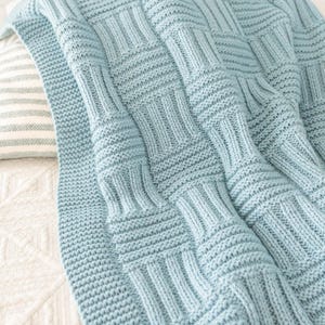 Può includere: Una coperta lavorata a maglia color azzurro con un motivo a intreccio testurizzato composto da sezioni a coste alternate verticalmente e orizzontalmente. Il tessuto presenta una tonalità blu tenue e uniforme, con un dettagliato motivo geometrico.