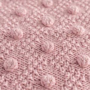 Pu&ograve; includere: Primo piano di una coperta lavorata a maglia in una delicata tonalit&agrave; rosa. La coperta presenta un motivo strutturato con pompon arrotondati in rilievo, distribuiti uniformemente sulla superficie. Le maglie creano un design dettagliato e tattile, perfetto per l'arredamento della casa.