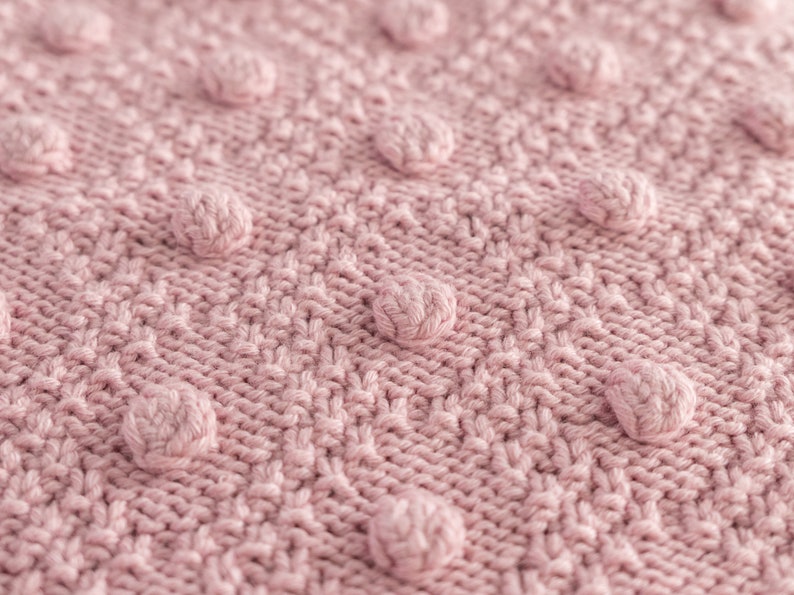 Pu&ograve; includere: Primo piano di una coperta lavorata a maglia in una delicata tonalit&agrave; rosa. La coperta presenta un motivo strutturato con pompon arrotondati in rilievo, distribuiti uniformemente sulla superficie. Le maglie creano un design dettagliato e tattile, perfetto per l'arredamento della casa.