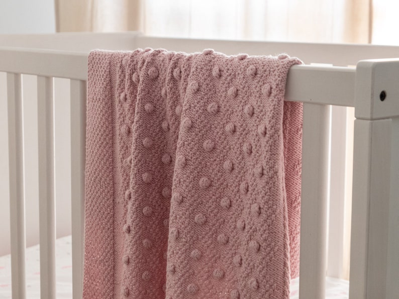 May include: A soft, pink knitted blanket with a textured bobble design, draped over a white crib. The blanket's pattern is visible, showcasing the detailed knitwork. The overall aesthetic is cozy and inviting, perfect for a nursery.