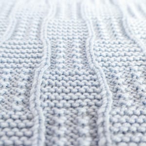 May include: Close-up of a light blue knitted fabric with a cable knit design. The texture is soft and the stitches are clearly visible, creating a cozy and textured appearance. The fabric is likely used for blankets or sweaters.
