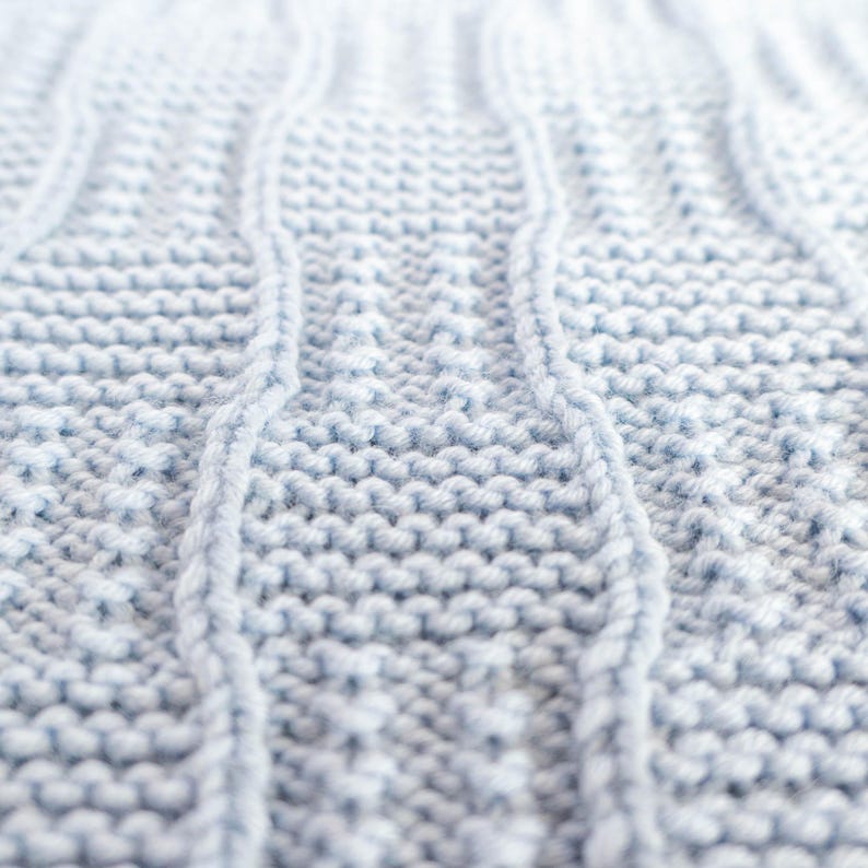 May include: Close-up of a light blue knitted fabric with a cable knit design. The texture is soft and the stitches are clearly visible, creating a cozy and textured appearance. The fabric is likely used for blankets or sweaters.