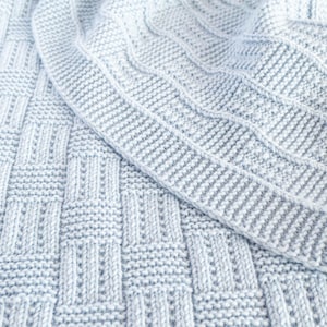 May include: A close-up of a light blue knitted blanket. The blanket features a textured pattern with a woven basket design and a ribbed border. The soft, cozy blanket is perfect for a baby or as a decorative throw.