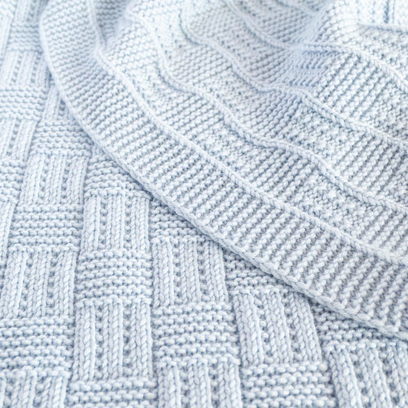 May include: A close-up of a light blue knitted blanket. The blanket features a textured pattern with a woven basket design and a ribbed border. The soft, cozy blanket is perfect for a baby or as a decorative throw.