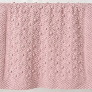 Pu&ograve; includere: Una morbida coperta rosa lavorata a maglia con un motivo a pois strutturato. La coperta &egrave; rettangolare e sembra essere fatta di un filato spesso. La coperta &egrave; esposta su un'asta di legno, suggerendo che si tratti di un oggetto decorativo.