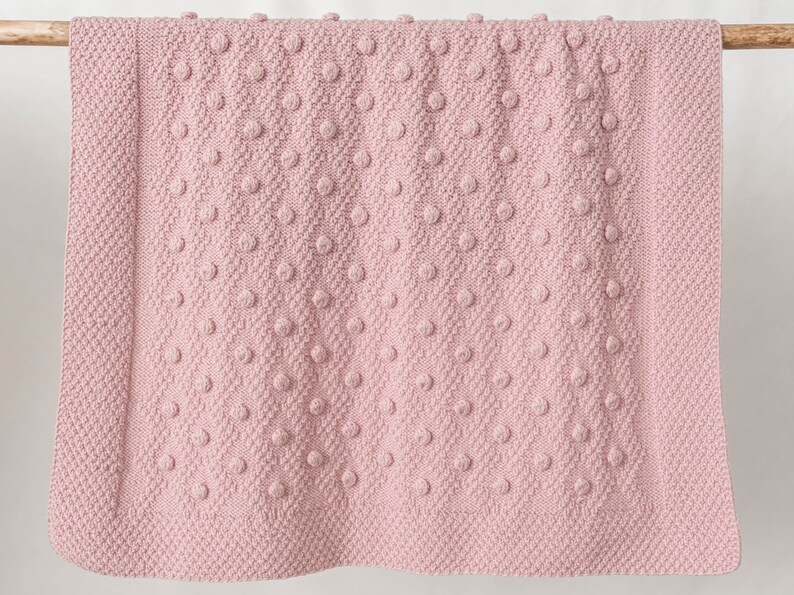 Pu&ograve; includere: Una morbida coperta rosa lavorata a maglia con un motivo a pois strutturato. La coperta &egrave; rettangolare e sembra essere fatta di un filato spesso. La coperta &egrave; esposta su un'asta di legno, suggerendo che si tratti di un oggetto decorativo.