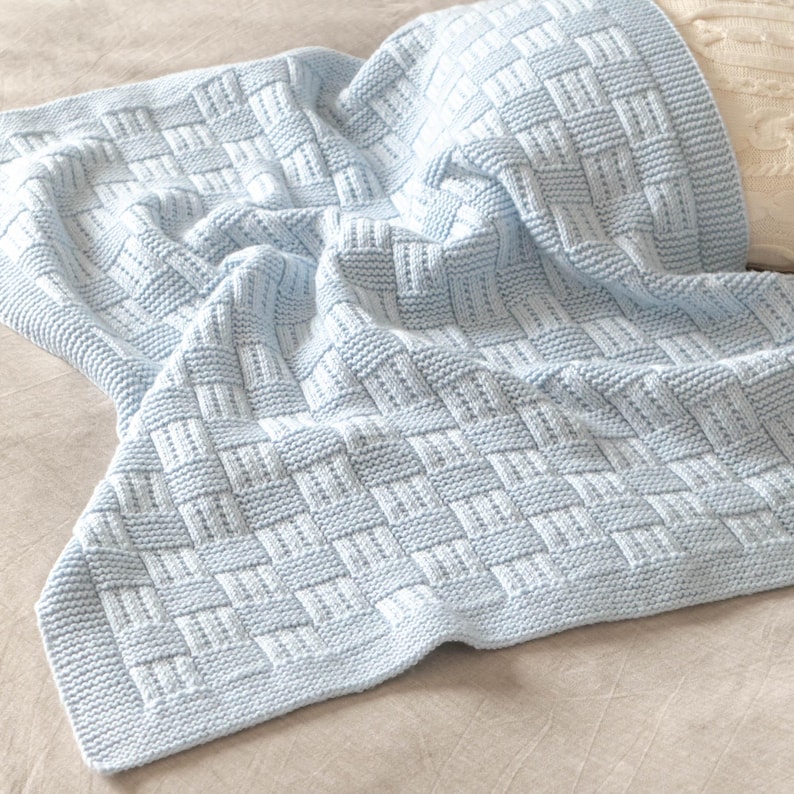 May include: A light blue knitted blanket with a woven basket weave pattern. The blanket is made of soft material and has a button closure. The blanket is folded over, showing the texture and design.