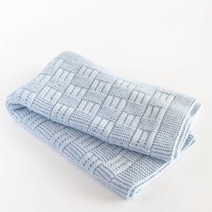 May include: A folded, light blue knitted blanket. The blanket features a woven basket weave pattern. The soft blanket is folded in half, showing the texture and design. The blanket is made of a soft material.