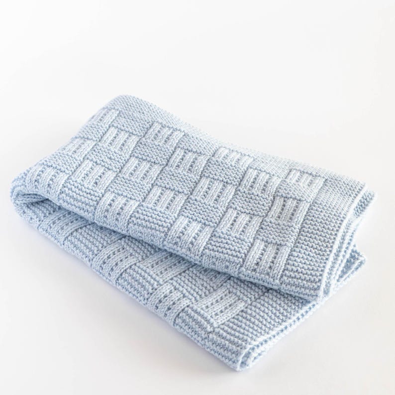 May include: A folded, light blue knitted blanket. The blanket features a woven basket weave pattern. The soft blanket is folded in half, showing the texture and design. The blanket is made of a soft material.