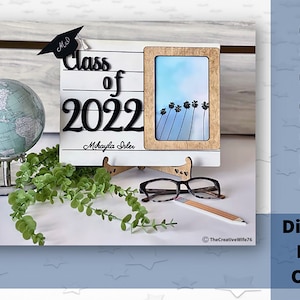 May include: White wooden sign with a black graduation cap, the text "Class of 2022" and the name "Mikayla Soles". A small picture frame with a blue sky and palm trees is attached to the sign. A globe, greenery, glasses, and a pencil are in the background.