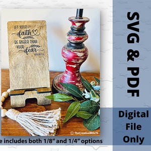 May include: A wooden phone stand with a laser-engraved inspirational quote: "Let your faith be bigger than your fear." The stand is made of brown wood and has a rustic finish. The quote is in black lettering and includes a small heart symbol. The stand is sitting on a wooden table with a decorative tassel and a red and black lamp.