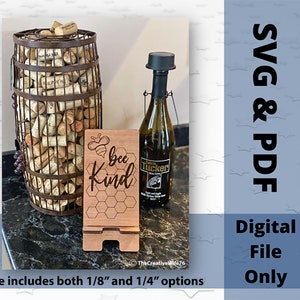 Könnte beinhalten: Ein Holzschild mit dem Text "bee kind" und einem Wabenmuster. Das Schild steht auf einem kleinen Holzständer. Das Schild steht vor einer Weinflasche und einem Fass voller Weinkorken.