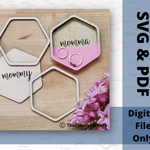 May include: Three hexagonal wooden trays, two with the word "mommy" and one with the word "momma". The tray with the word "momma" has a pink and gold glitter finish. The trays are on a wooden surface.