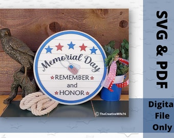 May include: A white round wooden sign with blue and red accents. The sign says "Memorial Day" with the words "Remember" and "Honor" in smaller text. The sign is decorated with stars and a small American flag.