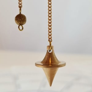 Puede incluir: Un péndulo de color dorado con una punta puntiaguda y un peso redondo y texturizado al final de la cadena.