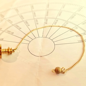 Puede incluir: Un péndulo dorado con un fondo redondo y lastrado y una cadena unida a una pequeña cuenta redonda. El péndulo descansa sobre un gráfico de papel blanco con un círculo y números.