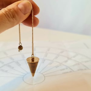 May include: A gold-colored pendulum with a conical shape and a spiral design hangs from a gold chain over a white chart with concentric circles.
