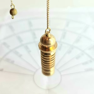 Puede incluir: Un péndulo de latón cuelga de una cadena, posicionado sobre un papel blanco con una cuadrícula semicircular. El péndulo tiene un cuerpo cilíndrico con estrías horizontales y una parte superior redondeada. Una pequeña esfera texturizada cuelga de una cadena separada arriba.