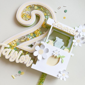 May include: A white and yellow cake topper featuring a number 2, the word "Taylor" and a lemonade stand with the word "Lemonade" on it. The number 2 is decorated with yellow and green sprinkles and lemon slices. The lemonade stand has a white roof with a yellow lemon on top and white daisies around it.