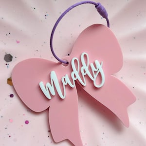 Può includere: Un fiocco rosa con il nome "Maddy" scritto in bianco. Il fiocco è attaccato a uno spago viola.