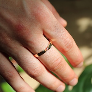 Pode incluir: Um anel dourado é usado em um dedo. O anel tem um design simples e polido. O anel é feito de um metal brilhante e reflexivo. O fundo está desfocado, com toques de folhagem verde e luz solar.