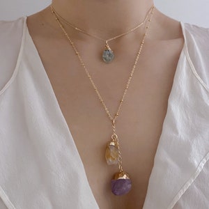 Può includere: Una collana a catena dorata con tre ciondoli di cristallo. Il ciondolo superiore è un cristallo blu chiaro, il ciondolo centrale è un cristallo giallo e il ciondolo inferiore è un cristallo viola.
