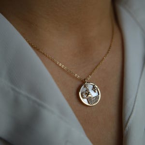 Könnte beinhalten: Eine goldfarbene Halskette mit einem kreisförmigen Anhänger. Der Anhänger hat einen weißen Hintergrund mit einem Design aus Stern, Mond und kleinen Steinen. Die Halskette wird über einem weißen Hemd getragen.