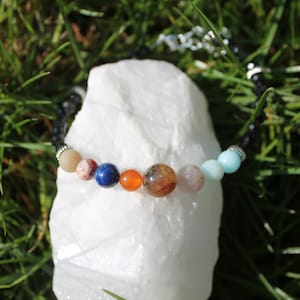 Könnte beinhalten: Ein Armband mit bunten Edelsteinperlen, die Planeten darstellen. Das Armband hat ein schwarzes Perlenband mit silberfarbenen Akzenten und einem Verschluss. Die Perlen sind in verschiedenen Größen und Farben, darunter Blau, Orange und Braun.