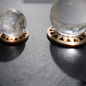 Könnte beinhalten: Zwei klare Glaskugeln, eine große und eine kleine, liegen auf runden, hellbraunen Holzuntersetzern. Die Untersetzer haben ein geometrisches Design. Die Kugeln scheinen aus Kristall oder Glas zu sein.