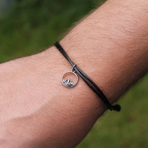 Könnte beinhalten: Ein schwarzes Kordelarmband mit einem silbernen Anhänger, der eine Bergsilhouette zeigt.