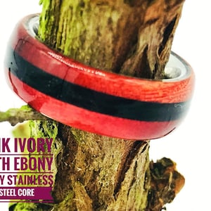 May include: A wooden ring with a pink ivory and ebony inlay. The ring has a stainless steel core. "PINK IVORY WITH EBONY INLAY STAINLESS STEEL CORE" is written on the image.