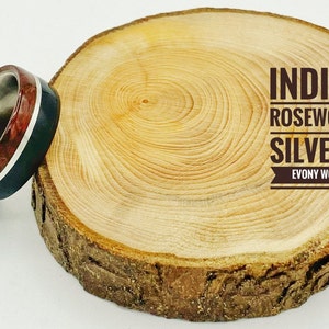 May include: A wooden ring with a silver band and a red and black inlay. The ring is on a wooden surface with the text "INDIAN ROSEWOOD, SILVER & EVONY WOOD".