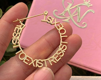 May include: A gold hoop earring featuring the repeating text "RESIST TO EXIST" integrated into the circular band design. The earring is held against a background of green leaves, a white box, and a pink box, both displaying a gold logo with a crown.
