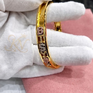 Pode incluir: Uma pulseira de bracelete dourada com as letras "BHSKY" escritas em pedras preciosas brilhantes. A pulseira tem um acabamento polido e é segurada por uma mão enluvada. As letras são numa mistura de cores, incluindo rosa e transparente.