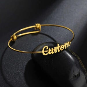May include: A gold-toned adjustable bangle bracelet with the word "Custom" engraved on it.