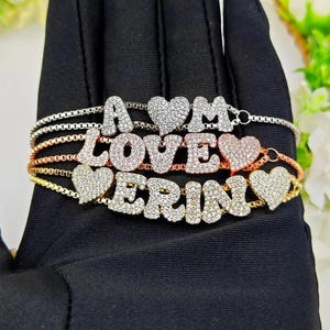 May include: Several personalized bracelets spelling out "A", "LOVE", and "ERIN" with clear gemstones. Each bracelet has a heart-shaped charm. The bracelets are displayed on a black surface.