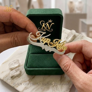 May include: A two-tone gold and silver name ring featuring the script text Kup Kake with a small heart accent. The ring is displayed in a dark green velvet jewelry box with the gold logo KN printed on the interior lid of the box.