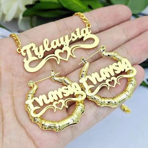 May include: Gold-tone necklace and earrings with the names "Tylaysia" and "Enarya" in a cursive font with a heart design. The earrings are bamboo-style hoops.