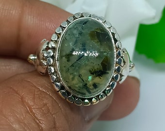 Könnte beinhalten: Ein silberner Ring mit einem ovalen, grün-schwarzen Edelstein, der in einer Perlenfassung gefasst ist. Der Ring hat ein dekoratives Design auf der Ringschiene.