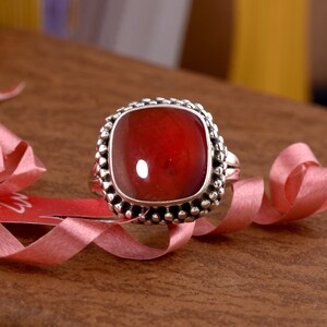 May include: A silver ring with a red gemstone set in a square bezel. The ring has a textured band and is surrounded by a row of small silver beads.
