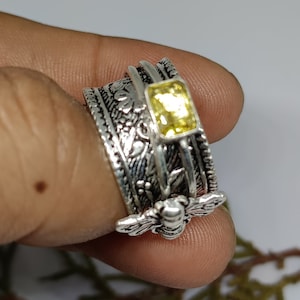 May include: A silver spinning ring with a yellow gemstone and a bee design. The ring has intricate floral details and is worn on a finger.