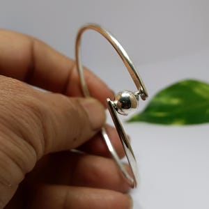 May include: A silver bangle bracelet featuring a smooth, round ball charm. The bracelet is open and has a simple, modern design.
